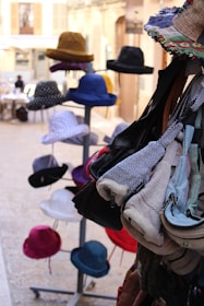 A collection of colorful hats displayed on stands and various fabric bags hanging on the right side. The setting appears to be an outdoor market or a shop with a cobblestone street in the background.