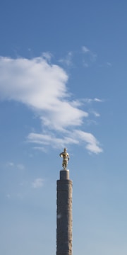 A golden statue stands atop a tall, intricately designed column against a clear blue sky with scattered clouds.