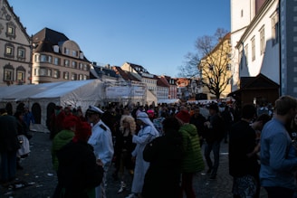 A lively outdoor gathering set in a town square, featuring crowds of people, some in costume. Historic European-style buildings line the square, with a large white tent set up as part of the event. The sky is clear and blue, and the lighting suggests a late afternoon setting. A variety of outfits and costumes, including some resembling uniforms and others more extravagant, hint at a festive occasion.