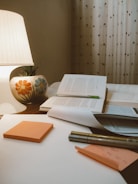 A cozy study setting features an open book with a green bookmark, placed on a wooden surface alongside several closed books. An illuminated lamp with a colorful floral design on its base casts a warm glow. In the foreground, orange sticky notes, a pen, and a sheet of paper with handwritten notes can be seen. The background is adorned with patterned curtains.