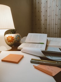A cozy study setting features an open book with a green bookmark, placed on a wooden surface alongside several closed books. An illuminated lamp with a colorful floral design on its base casts a warm glow. In the foreground, orange sticky notes, a pen, and a sheet of paper with handwritten notes can be seen. The background is adorned with patterned curtains.