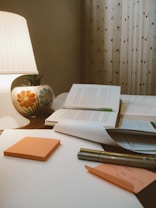 A cozy study setting features an open book with a green bookmark, placed on a wooden surface alongside several closed books. An illuminated lamp with a colorful floral design on its base casts a warm glow. In the foreground, orange sticky notes, a pen, and a sheet of paper with handwritten notes can be seen. The background is adorned with patterned curtains.