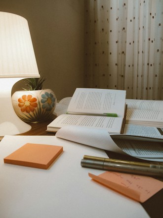 A cozy study setting features an open book with a green bookmark, placed on a wooden surface alongside several closed books. An illuminated lamp with a colorful floral design on its base casts a warm glow. In the foreground, orange sticky notes, a pen, and a sheet of paper with handwritten notes can be seen. The background is adorned with patterned curtains.