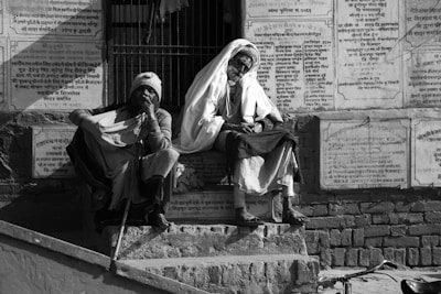 Two elderly individuals are sitting on stone steps in front of a wall covered in plaques with Hindi script. One wears a beanie and wraps a cloth around themselves, resting a hand on a walking stick. The other is in a white head covering and shawl, appearing introspective. The setting is outdoors, possibly near a religious or historical site.