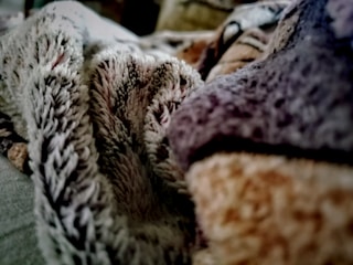 A close-up view of a textured fabric, possibly a blanket or plush surface, with a mix of colors including gray, brown, and hints of red. The fabric appears soft and fuzzy, with a focus on its detailed texture.