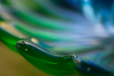 A close-up of a glass sculpture featuring smooth, flowing curves with a glossy surface. The colors transition seamlessly from green to blue, creating a mesmerizing effect.