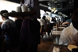 A busy café or restaurant with people sitting at tables and looking out of large windows. The lighting is dim, with natural light streaming in from the windows, creating a contrast with the darker interior. A server is carrying a tray with cups, and there are stylish overhead lamps adding to the cozy ambiance.