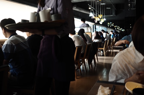A busy café or restaurant with people sitting at tables and looking out of large windows. The lighting is dim, with natural light streaming in from the windows, creating a contrast with the darker interior. A server is carrying a tray with cups, and there are stylish overhead lamps adding to the cozy ambiance.