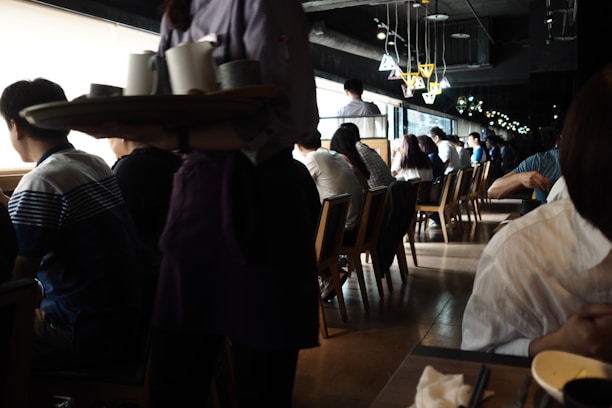 A busy café or restaurant with people sitting at tables and looking out of large windows. The lighting is dim, with natural light streaming in from the windows, creating a contrast with the darker interior. A server is carrying a tray with cups, and there are stylish overhead lamps adding to the cozy ambiance.