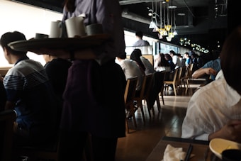 A busy café or restaurant with people sitting at tables and looking out of large windows. The lighting is dim, with natural light streaming in from the windows, creating a contrast with the darker interior. A server is carrying a tray with cups, and there are stylish overhead lamps adding to the cozy ambiance.