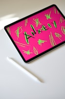 A tablet displaying the word 'Anxiety' in bold letters with a bright pink background and neon green scribbles. The tablet is placed on a white surface next to a white stylus pen.