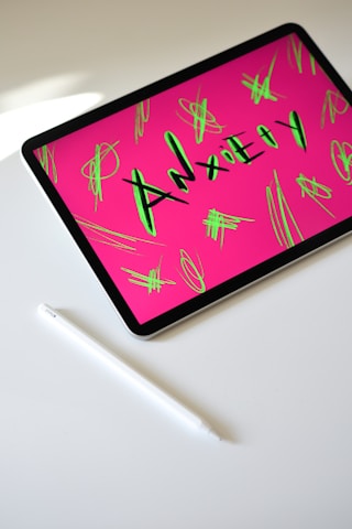 A tablet displaying the word 'Anxiety' in bold letters with a bright pink background and neon green scribbles. The tablet is placed on a white surface next to a white stylus pen.