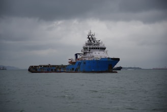 A large, blue cargo or utility ship is floating on a calm sea under a cloudy, overcast sky. The vessel is weathered and industrial-looking, with various equipment and structures on its deck. It is positioned slightly to the right of the center with land visible in the distant background.