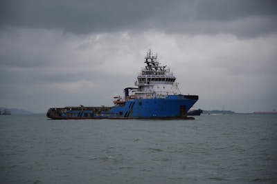 A large, blue cargo or utility ship is floating on a calm sea under a cloudy, overcast sky. The vessel is weathered and industrial-looking, with various equipment and structures on its deck. It is positioned slightly to the right of the center with land visible in the distant background.