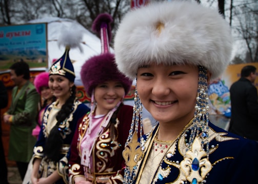 Several people wearing traditional, colorful costumes with intricate embroidery and fur hats are gathered at an outdoor event. They are smiling and appear to be part of a cultural celebration or festival. In the background, there are banners and tents, indicating a festive atmosphere.