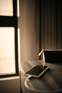 A smartphone lies on a wooden table, connected to a charging cable. Soft ambient light filters through a window, casting a warm glow on the surroundings. In the background, curtains add a sense of calm and coziness to the scene.