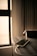 A smartphone lies on a wooden table, connected to a charging cable. Soft ambient light filters through a window, casting a warm glow on the surroundings. In the background, curtains add a sense of calm and coziness to the scene.