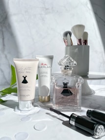 A selection of beauty products is arranged on a marble surface. On the left, two tubes of lotion are displayed, both featuring elegant cursive branding. In the center, an intricately designed perfume bottle with a stylish dress motif is prominently featured. To the right, makeup brushes with shiny metallic handles stand upright in a sleek holder. A black mascara tube lies in the foreground. The composition is accentuated by soft natural light and subtle shadows.