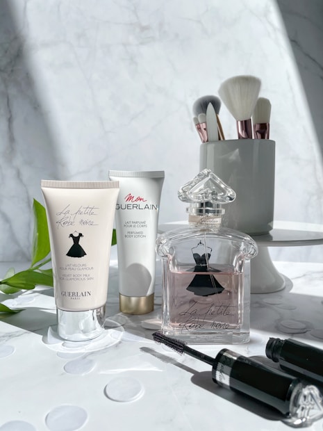 A selection of beauty products is arranged on a marble surface. On the left, two tubes of lotion are displayed, both featuring elegant cursive branding. In the center, an intricately designed perfume bottle with a stylish dress motif is prominently featured. To the right, makeup brushes with shiny metallic handles stand upright in a sleek holder. A black mascara tube lies in the foreground. The composition is accentuated by soft natural light and subtle shadows.