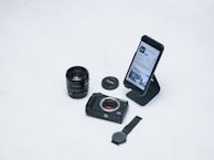 A flat lay of photography-related items including a camera lens, a camera body, a lens cap, a smartphone displaying a photography app, and a watch. The smartphone is placed on a stand and the overall arrangement is on a white surface.