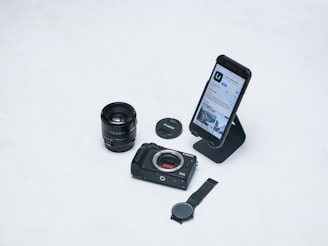 A flat lay of photography-related items including a camera lens, a camera body, a lens cap, a smartphone displaying a photography app, and a watch. The smartphone is placed on a stand and the overall arrangement is on a white surface.