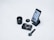 A flat lay of photography-related items including a camera lens, a camera body, a lens cap, a smartphone displaying a photography app, and a watch. The smartphone is placed on a stand and the overall arrangement is on a white surface.
