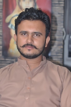 A man with a well-groomed beard and styled hair is wearing a traditional brown kurta. The background features a blurred painting that seems to depict a person.