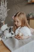 A young girl with long, light brown hair is gently holding a white rabbit and an egg, sitting beside another white and black rabbit on a soft gray fabric. She is in a cozy setting with a vase of white pussy willow branches in the background.