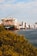 A large cargo ship with stacked colorful containers is docked near an urban area with tall buildings. In the foreground, there is dense greenery bordering a body of water.
