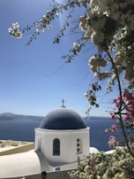 A picturesque scene featuring a classic Greek Orthodox church with a blue dome and white walls overlooking the deep blue sea. Bougainvillea flowers in shades of pink and white cascade in the foreground. The clear sky and bright sunlight create a serene atmosphere.