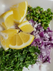 Sliced yellow lemons, chopped green chili peppers, diced red onions, and finely chopped fresh cilantro arranged on a white surface.