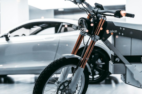 A close-up of a modern electric motorcycle with sleek design is in the foreground. Behind it, a silver luxury car with smooth curves is parked indoors. The setting is likely a showroom, and the lighting casts a reflective sheen on both vehicles, highlighting their contemporary features.