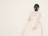 A person with dark hair styled in a bun adorned with decorative hairpieces is wearing a long-sleeved, light-colored gown. The gown has a cinched waist and a full skirt with a sheer overlay. The background is plain, enhancing the elegant and minimalist aesthetic.