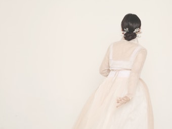 A person with dark hair styled in a bun adorned with decorative hairpieces is wearing a long-sleeved, light-colored gown. The gown has a cinched waist and a full skirt with a sheer overlay. The background is plain, enhancing the elegant and minimalist aesthetic.