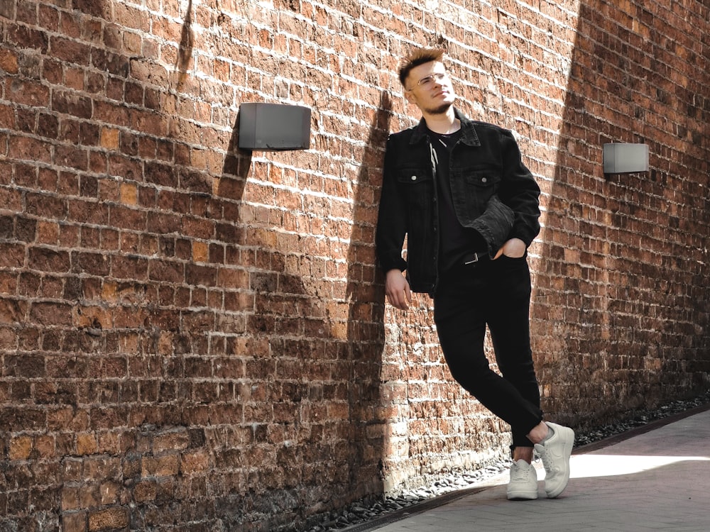 man in black leather jacket and black pants leaning on brown brick wall