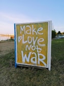 A brightly colored sign with a yellow background displays the phrase 'Make Love Not War' in large white letters. The sign is placed outdoors on a grassy area with a clear blue sky above and some trees in the background.