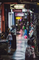 A nighttime scene in a narrow alleyway with various shops on either side. A person wearing a headscarf is seen holding a shopping bag and examining goods at a street vendor's stall. The ground appears wet, reflecting colorful lights from the surroundings. Signs indicating a food court and other shops hang above.