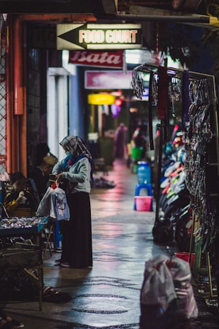 A nighttime scene in a narrow alleyway with various shops on either side. A person wearing a headscarf is seen holding a shopping bag and examining goods at a street vendor's stall. The ground appears wet, reflecting colorful lights from the surroundings. Signs indicating a food court and other shops hang above.