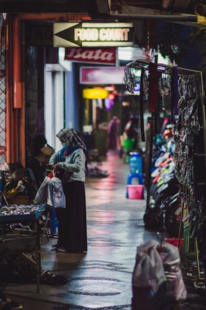 A nighttime scene in a narrow alleyway with various shops on either side. A person wearing a headscarf is seen holding a shopping bag and examining goods at a street vendor's stall. The ground appears wet, reflecting colorful lights from the surroundings. Signs indicating a food court and other shops hang above.
