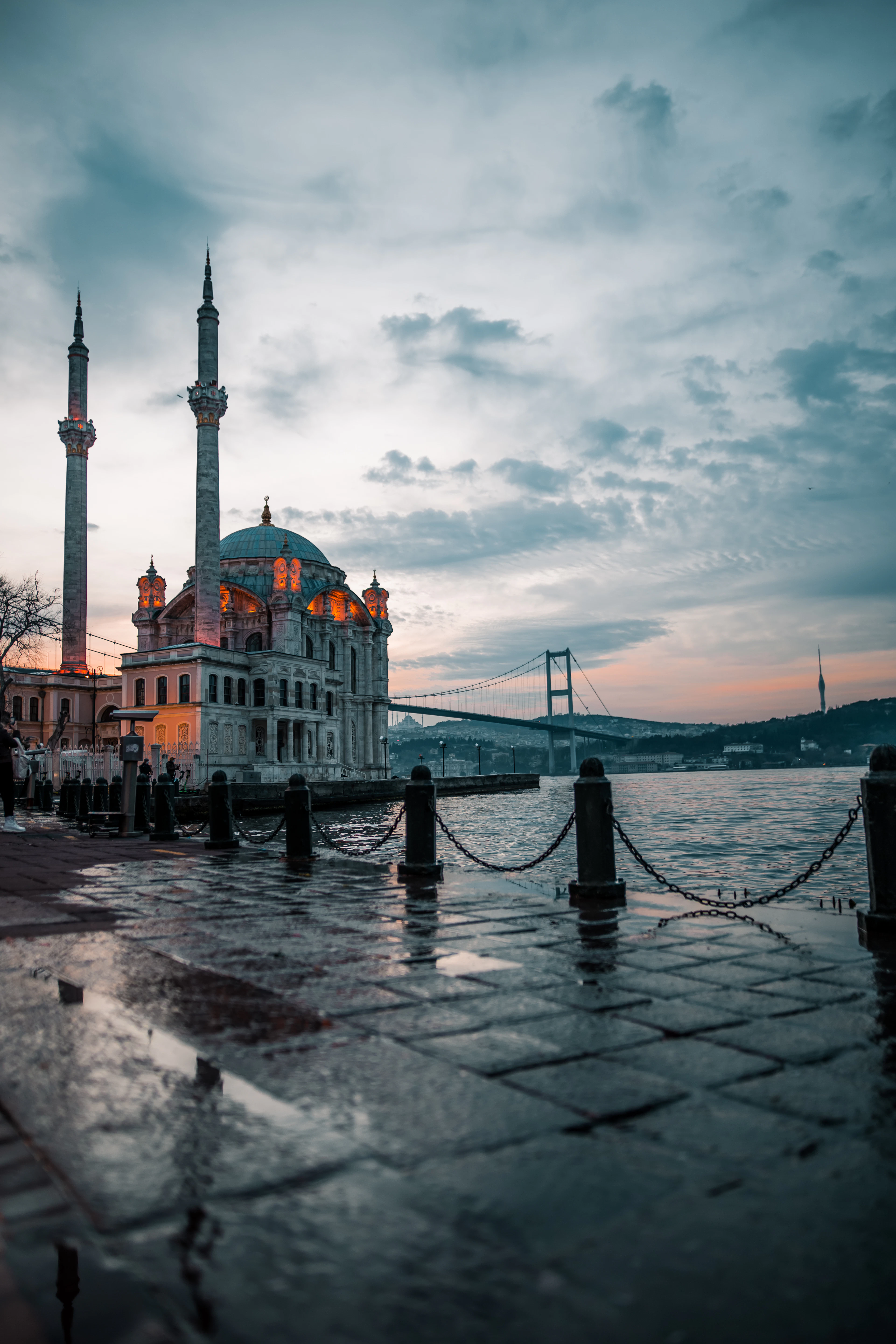 Ortakoy Mosque glows at dusk beside the Bosphorus Bridge, its lights reflected on rain-wet pavement.