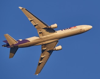 A commercial cargo airplane in flight against a clear blue sky. The aircraft is viewed from below and is painted in light colors with purple accents.