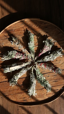 Herbs bundled with different colored strings are arranged in a circular pattern on a rustic wooden surface, casting shadows that enhance the texture of the wood.