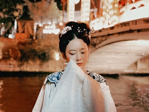 A woman in traditional attire stands by a softly lit riverside. Her hair is elegantly styled with ornamental hairpins and flowers. She holds a fan or cloth partially covering her face, adding an air of mystery. The background features a bridge and blurred lights reflecting on the water, creating a warm, serene ambiance.