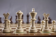 A close-up view of a chessboard with a focus on the white chess pieces, including the bishop, queen, king, and knight. The pieces are arranged prominently in the foreground, standing on a classic brown and beige checkered board.