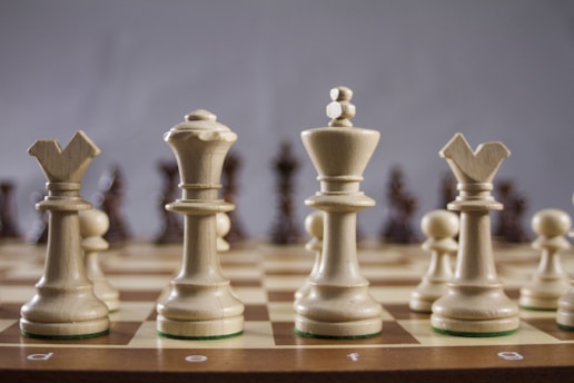 A close-up view of a chessboard with a focus on the white chess pieces, including the bishop, queen, king, and knight. The pieces are arranged prominently in the foreground, standing on a classic brown and beige checkered board.