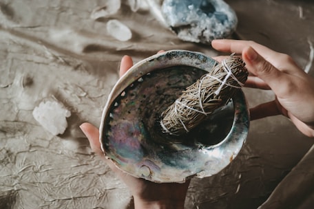 Hands hold a polished abalone shell containing a bundle of dried herbs wrapped with string. The surface is iridescent with dark and light hues, adding texture to the scene. Nearby, crystals rest on a rustic, fabric-covered background.