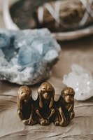 A bronze statue of the three wise monkeys, symbolizing 'see no evil, hear no evil, speak no evil,' is prominently featured in the foreground. The background includes blurred images of blue and white crystals, creating a serene and mystical ambiance.
