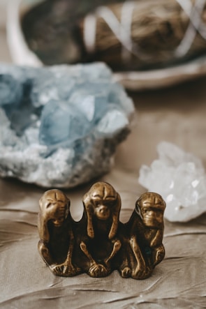 A bronze statue of the three wise monkeys, symbolizing 'see no evil, hear no evil, speak no evil,' is prominently featured in the foreground. The background includes blurred images of blue and white crystals, creating a serene and mystical ambiance.