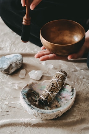A person is seen holding a wooden singing bowl with a mallet positioned over it, suggesting it is ready to be used. On a textured surface below are various objects including a bundle of sage tied with string, an abalone shell, and a few crystals or stones, creating a serene and spiritual atmosphere.