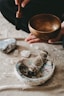 A person is seen holding a wooden singing bowl with a mallet positioned over it, suggesting it is ready to be used. On a textured surface below are various objects including a bundle of sage tied with string, an abalone shell, and a few crystals or stones, creating a serene and spiritual atmosphere.